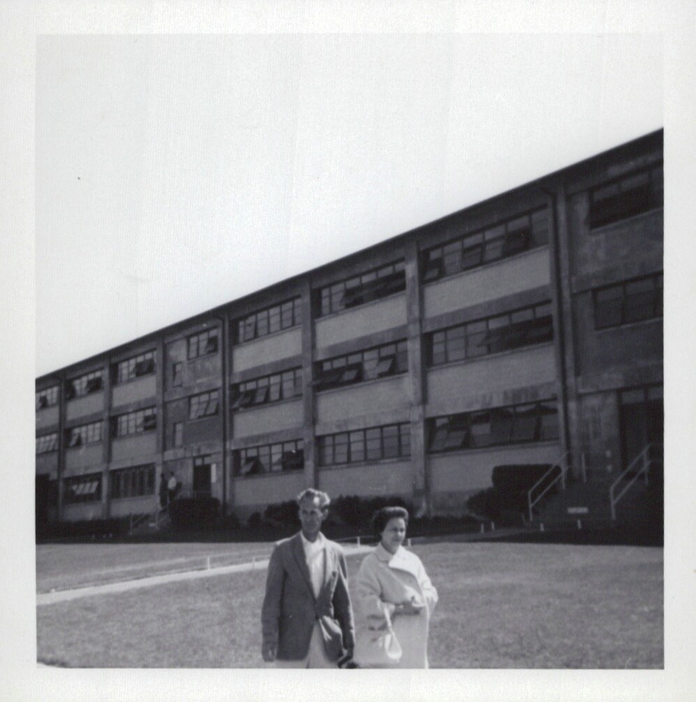 Two adults — a man in a suit and a woman in a coat — standing on a lawn in front of a large multi-storey institutional building with horizontal ribbon windows, black-and-white snapshot.