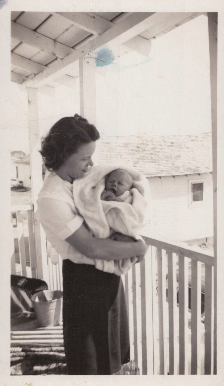A dark-haired woman in a blouse and skirt stands on a white-railed porch, cradling a swaddled newborn infant in her arms, in a mid-20th-century black-and-white snapshot.