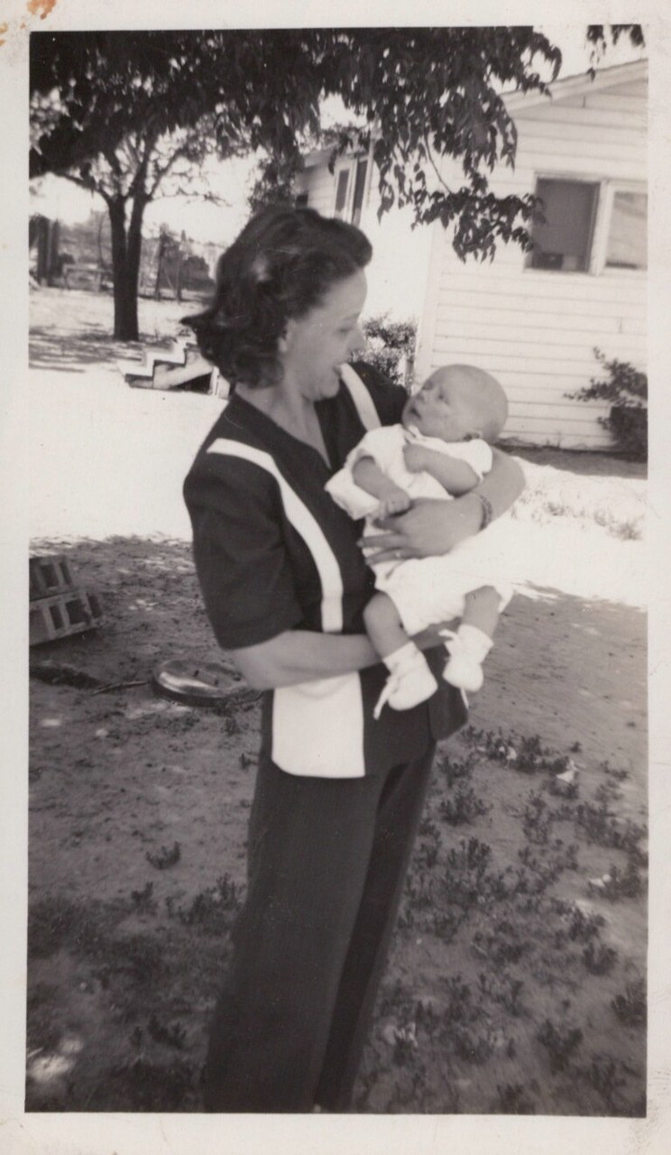 A woman in a dark top with white trim stands in a backyard in profile, cradling a newborn infant and looking down at the baby; black-and-white snapshot, circa 1940s–1950s