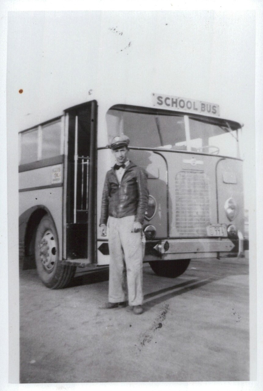 A young man in a leather jacket, bow tie, and flat cap stands in front of an old-style school bus with its door open, in a black-and-white photograph.