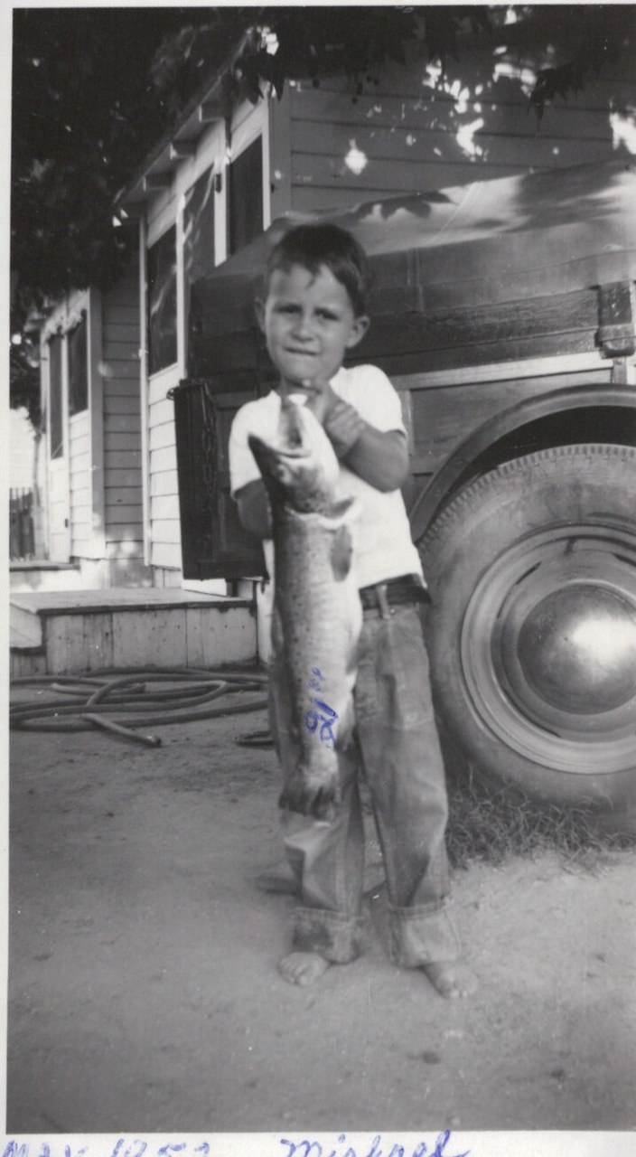 A barefoot young boy proudly holds a large fish almost as tall as himself, standing in a dirt yard beside an old truck, with a wood-sided house in the background; black-and-white photo with a handwritten caption reading a date and "Michael."