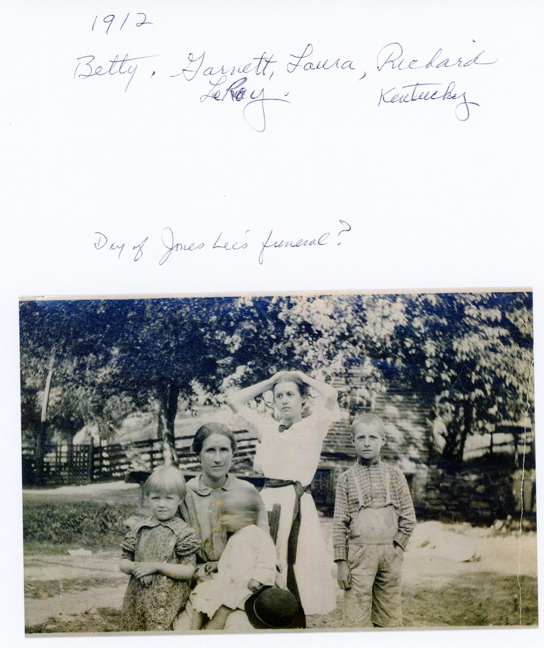 Four children posed outdoors in front of large trees and wooden fence, early 20th century.