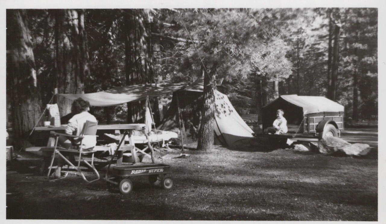 Black-and-white photo of a forested campsite with two people, canvas tents, a tarp canopy, a Radio Flyer wagon, and a vintage camping trailer among tall conifers
