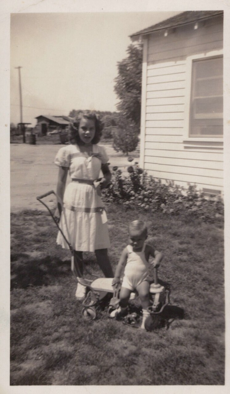 Black-and-white photo of an older girl in a belted dress standing on a lawn holding a child's wagon, with a toddler beside a small ride-on toy, next to a white clapboard house