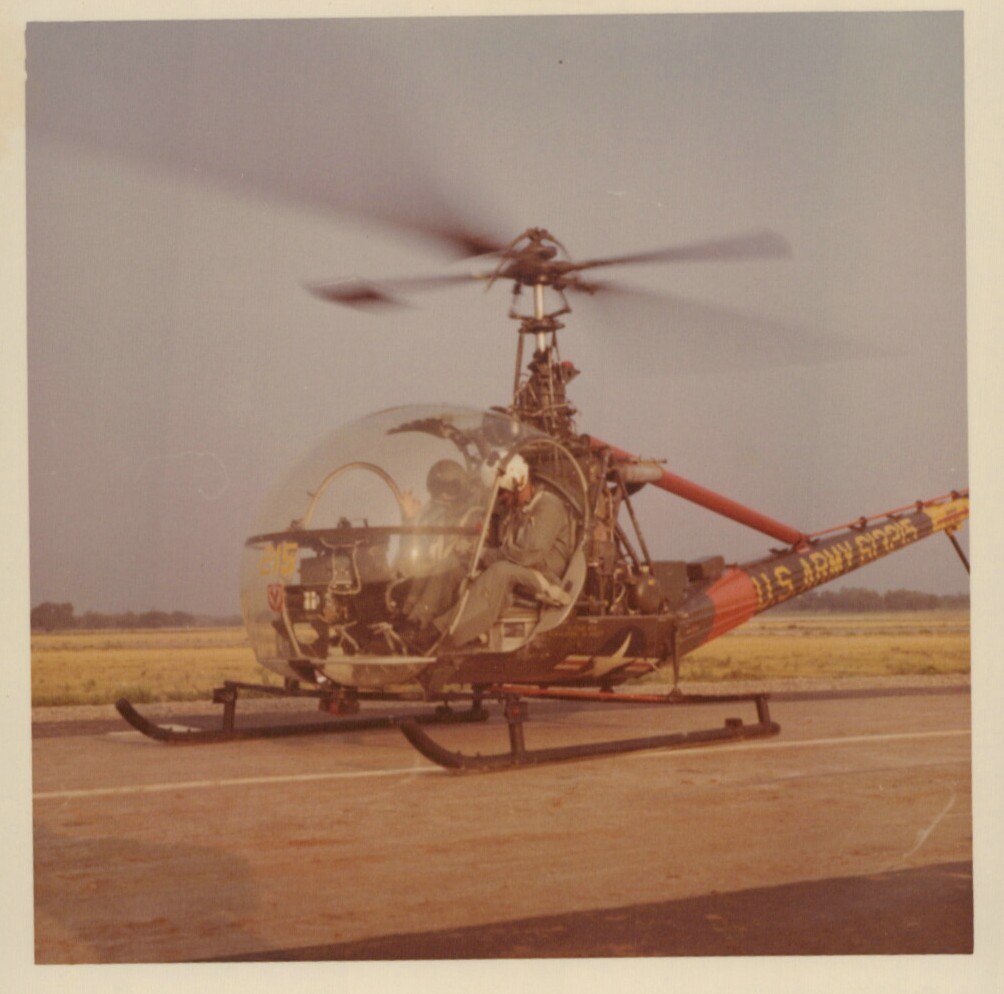 A U.S. Army Bell H-13-style bubble-canopy helicopter with rotor spinning on a military airfield tarmac, with a helmeted occupant visible in the cockpit and "U.S. ARMY" text on the red tail boom.