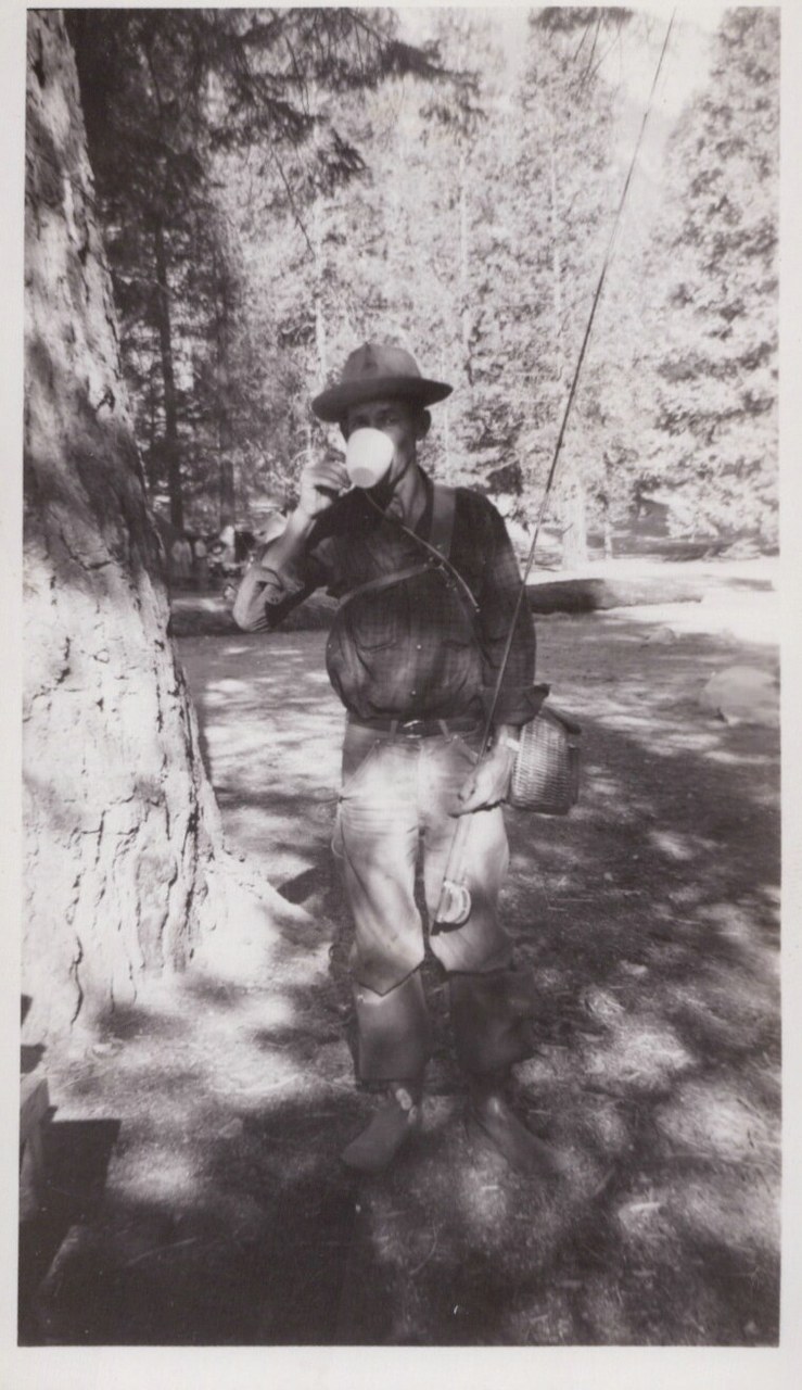 A person in a wide-brimmed hat, dark shirt, light trousers, and leather chaps drinks from a cup beside a large tree in a pine forest, carrying fishing gear including a wicker creel and rod