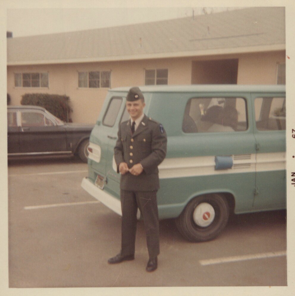 A young man in a U.S. Army dress uniform stands smiling in a parking lot in front of a teal-and-white van, with a low military-style building in the background; photo dated January 1967.
