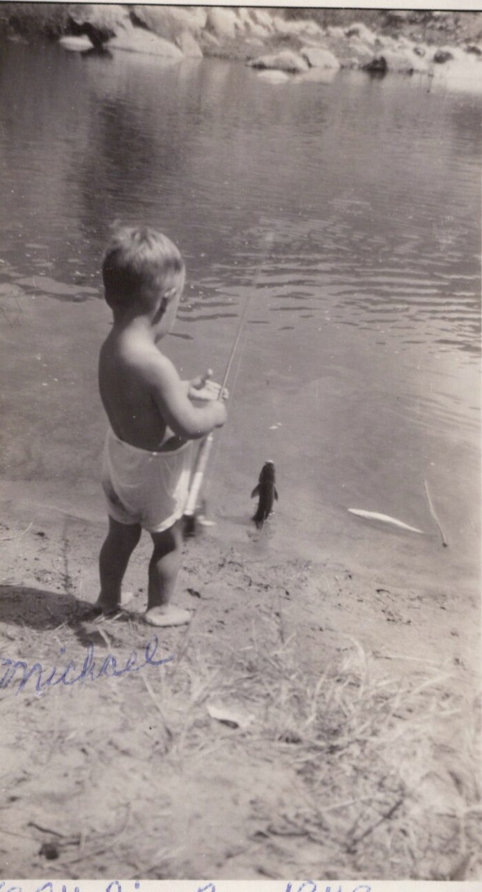 A shirtless toddler holding a fishing pole stands on a sandy riverbank with a small caught fish on the ground before them; black-and-white photo inscribed "Michael" in cursive