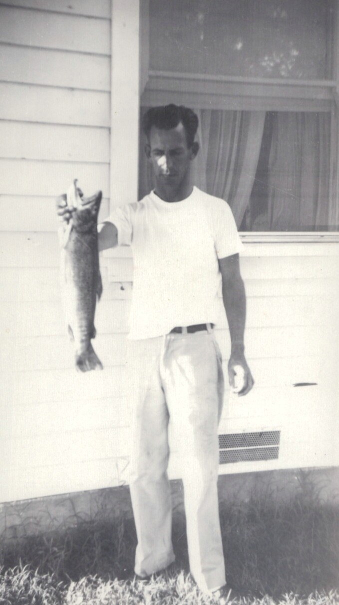 A man in a white T-shirt and light trousers stands on a lawn in front of a white clapboard house, holding up a large fish with one hand in a black-and-white snapshot.