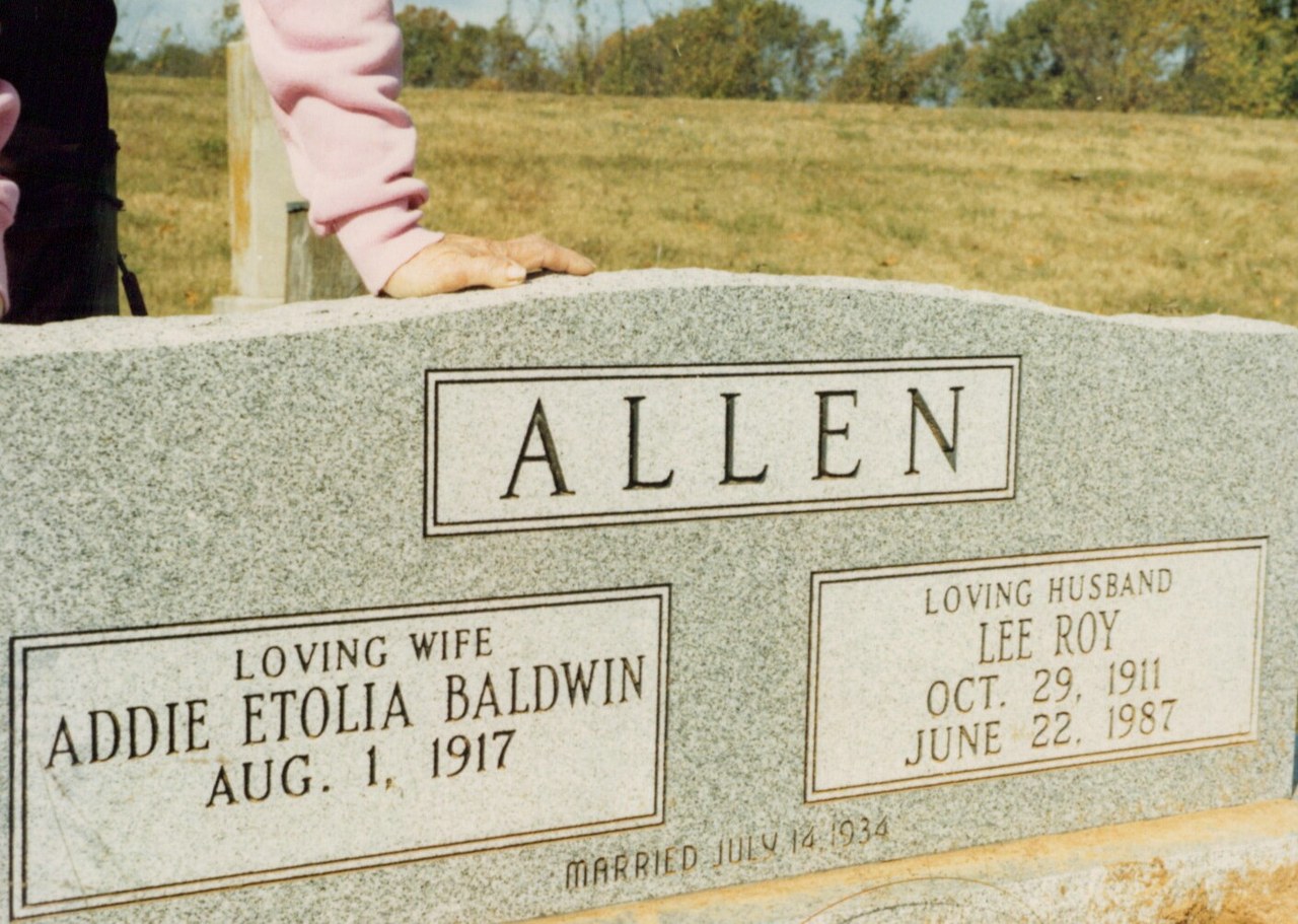 Grey granite double headstone for Lee Roy Allen (1911–1987) and Addie Etolia Baldwin Allen (b. 1917), married July 14, 1934, with a person's hand resting on top of the stone in a rural cemetery.