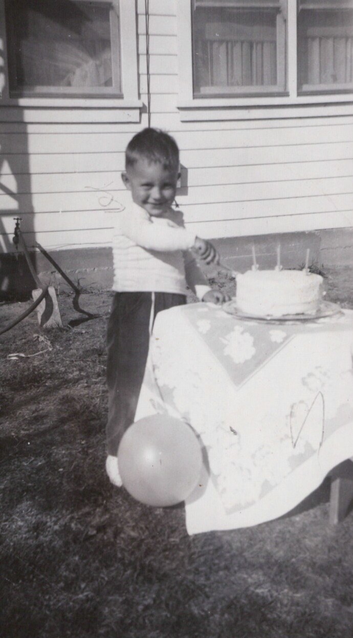 Young child at outdoor table with cake and balloon, white-sided house in background