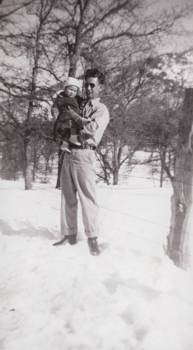 A man in sunglasses and cuffed trousers holds a bundled infant outdoors in a snowy rural setting, with bare trees and a barbed-wire fence in the background, in a vintage black-and-white photo.