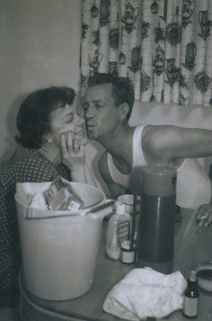 A smiling woman being kissed on the cheek by a man in a tank top, seated near a table with household items, black-and-white photo, circa 1950s.