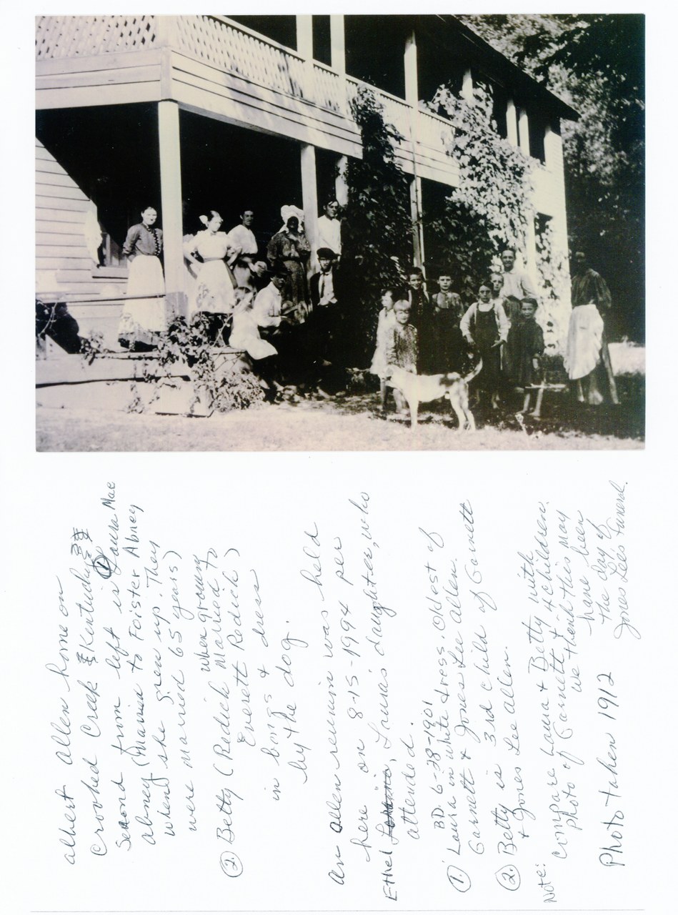 A group of people of mixed ages gathered outside a white wooden house with ivy and vegetation, circa early 20th century.