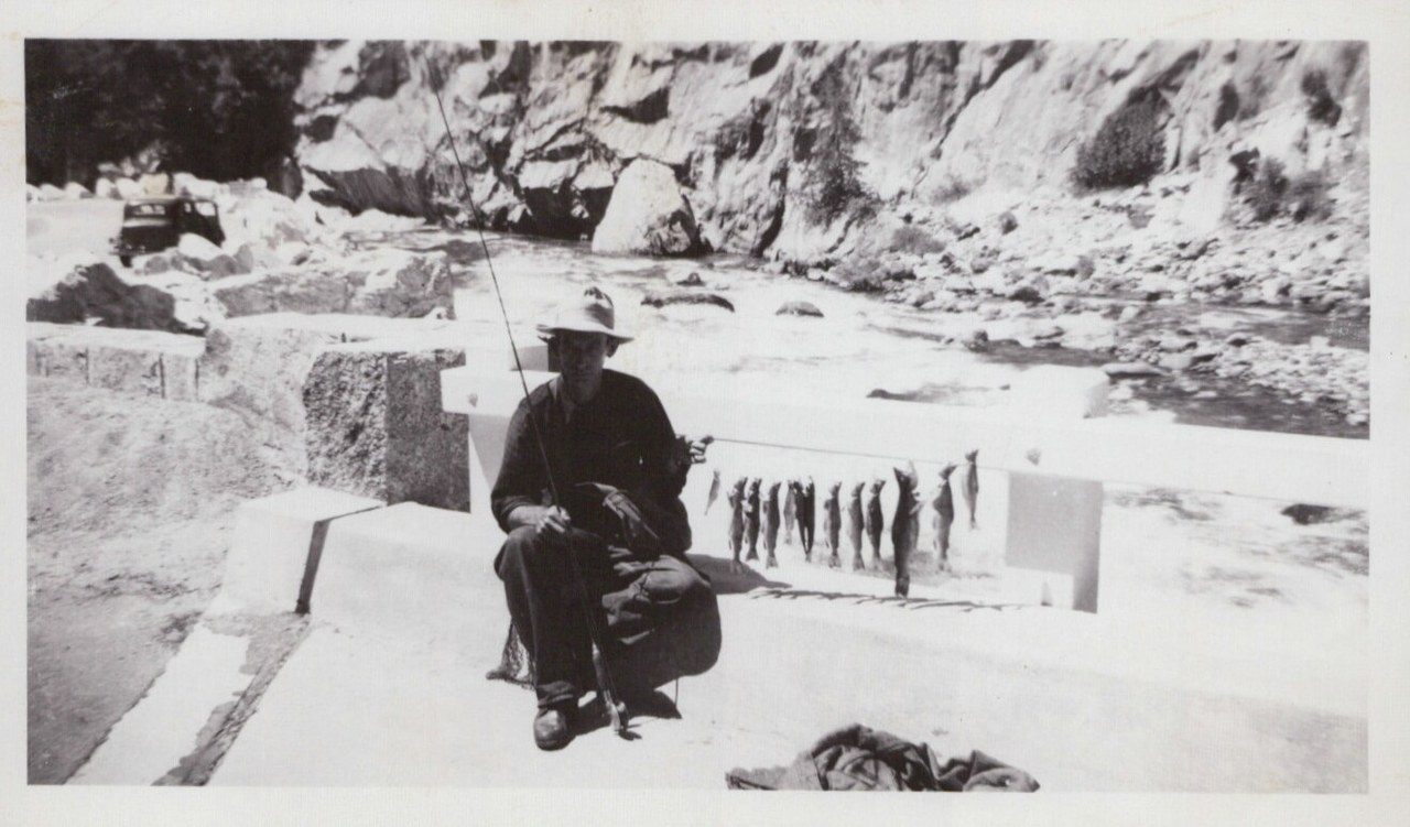 A person in a wide-brimmed hat sits on a concrete ledge holding a fishing rod beside a row of caught fish, with a rocky canyon river in the background, black-and-white snapshot.
