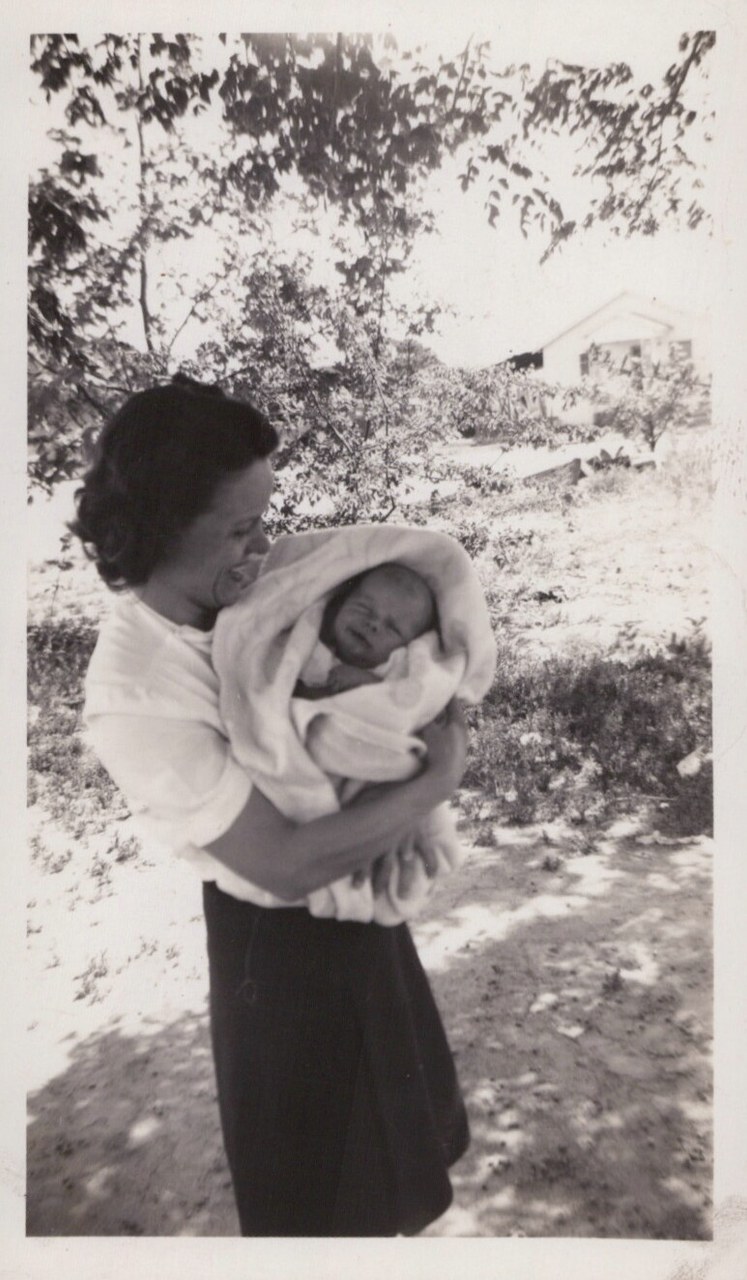 A woman with dark wavy hair cradles a swaddled newborn outdoors in a black-and-white snapshot, with trees and a small white building in the background.