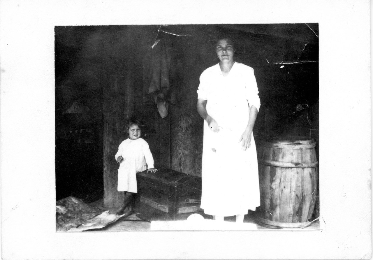 Black-and-white photograph of a woman in a white dress standing with a small child in a rustic interior with wooden posts and a barrel.