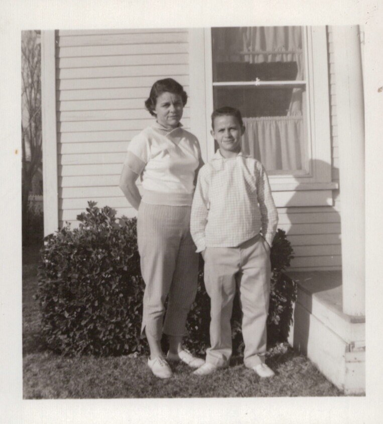Black-and-white snapshot of an adult woman and a young boy standing together on a lawn in front of a white clapboard house, likely late 1950s to early 1960s.