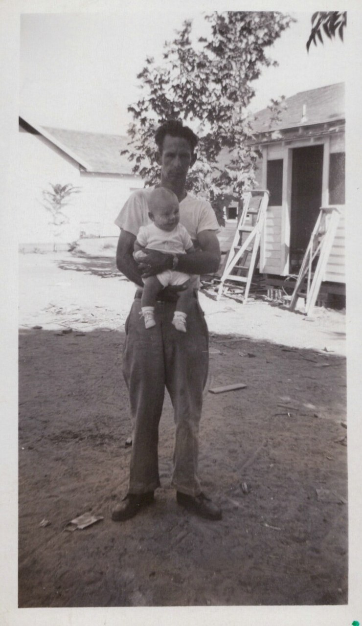 A man in a white T-shirt and high-waisted trousers holds a small infant outdoors in front of a wood-sided house with a ladder leaning against it, black-and-white photo circa 1940s–1950s