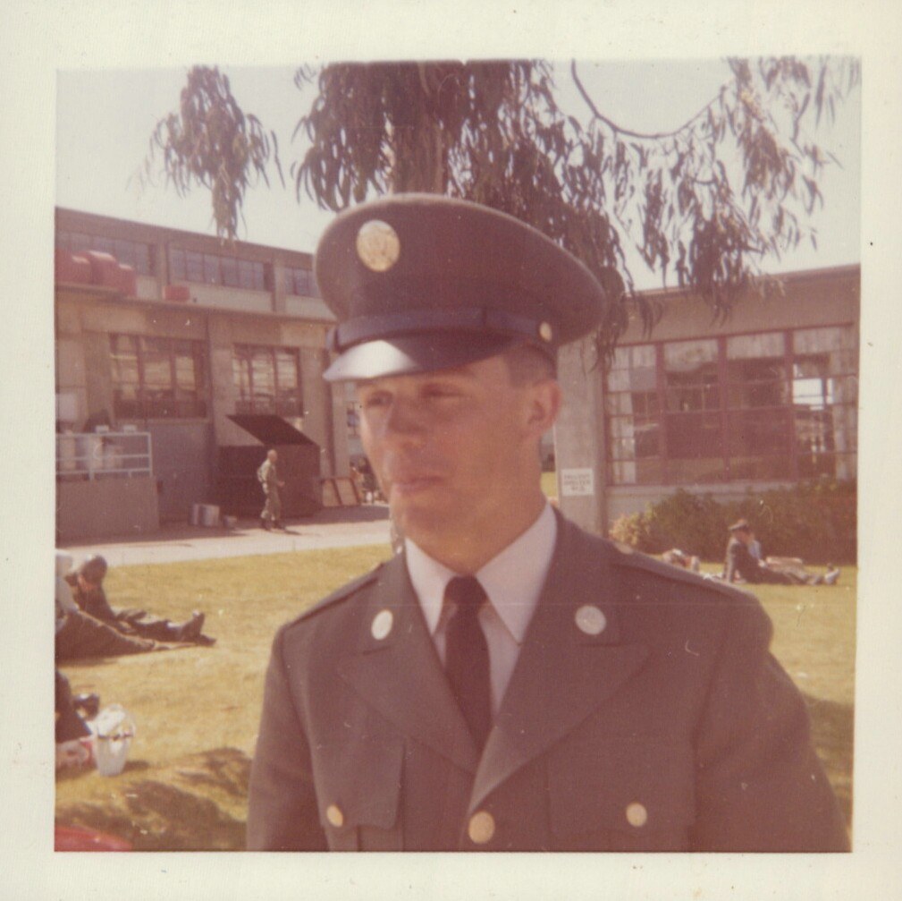 A young man in a U.S. Army Class A dress uniform and service cap stands on a grassy area in front of a large institutional building, with other uniformed figures visible in the background.