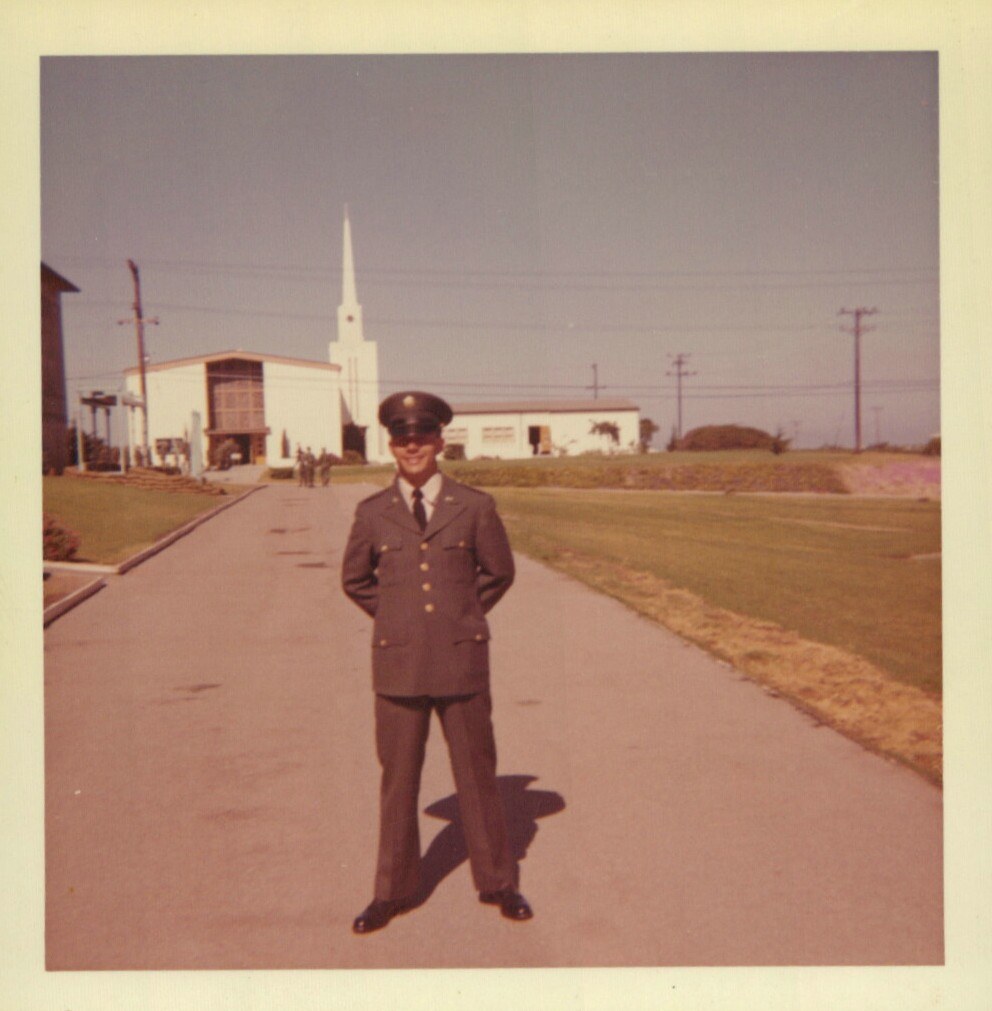 A smiling man in a dark olive-green U.S. military dress uniform and peaked cap stands on a paved path in front of a white chapel with a tall steeple on what appears to be a military base.