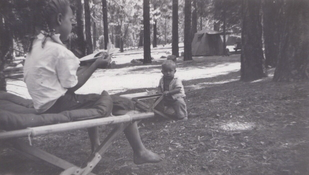 Two children at a forested campsite: one sits on a folding camp cot barefoot, the other is a toddler crouching on the ground; a canvas tent is visible among tall pine trees in the background.