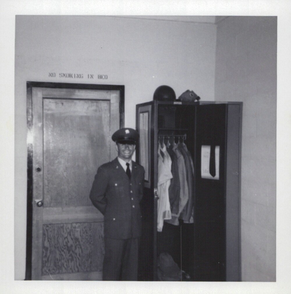 A smiling young man in a dark military dress uniform and garrison cap stands beside an open wall locker in a barracks room; a "No Smoking in Bed" sign is visible above the door to his left.