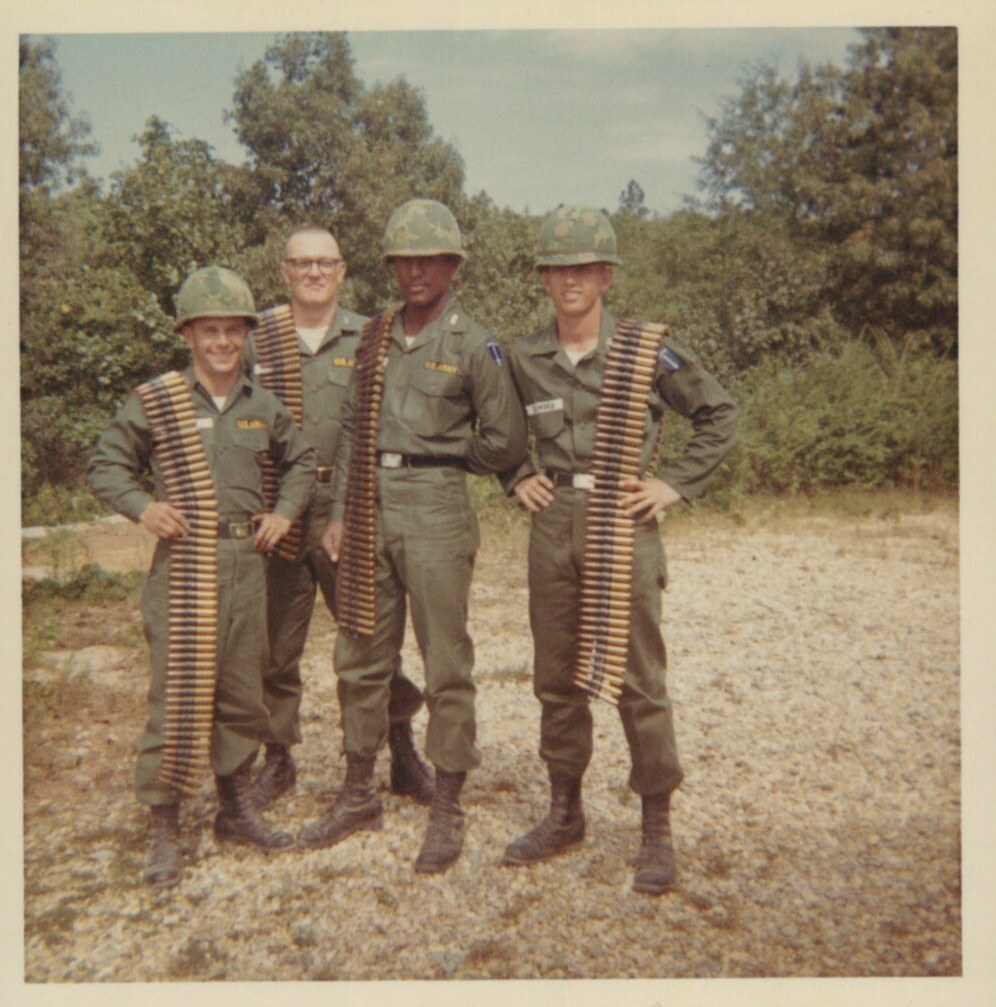 Four smiling U.S. Army soldiers in olive-drab uniforms and steel helmets pose outdoors, three of them draped with belts of linked ammunition, trees visible in background.