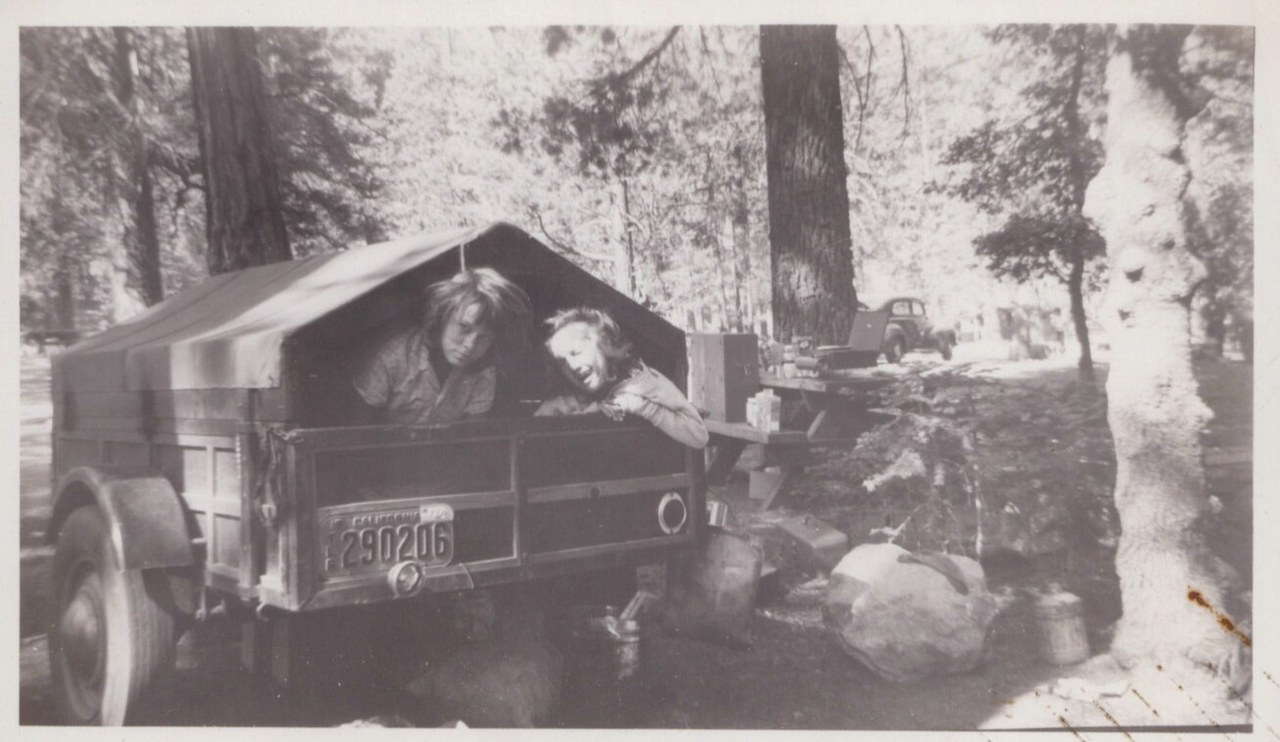 Two women lean out of a small canvas-roofed camping trailer with a California license plate (290206), parked at a wooded campsite; vintage car visible in background, black-and-white photo