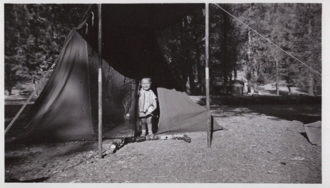 A smiling toddler stands at the entrance of a large canvas camping tent in a pine-forested campground, with small toy vehicles on the dirt ground in front of them