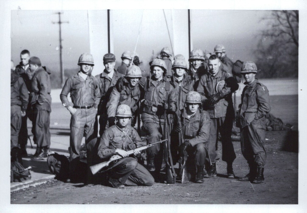 Group portrait of roughly 15–20 uniformed military soldiers in helmets and fatigues, posed outdoors on a military base, two holding rifles, black-and-white photo circa 1950s–1960s
