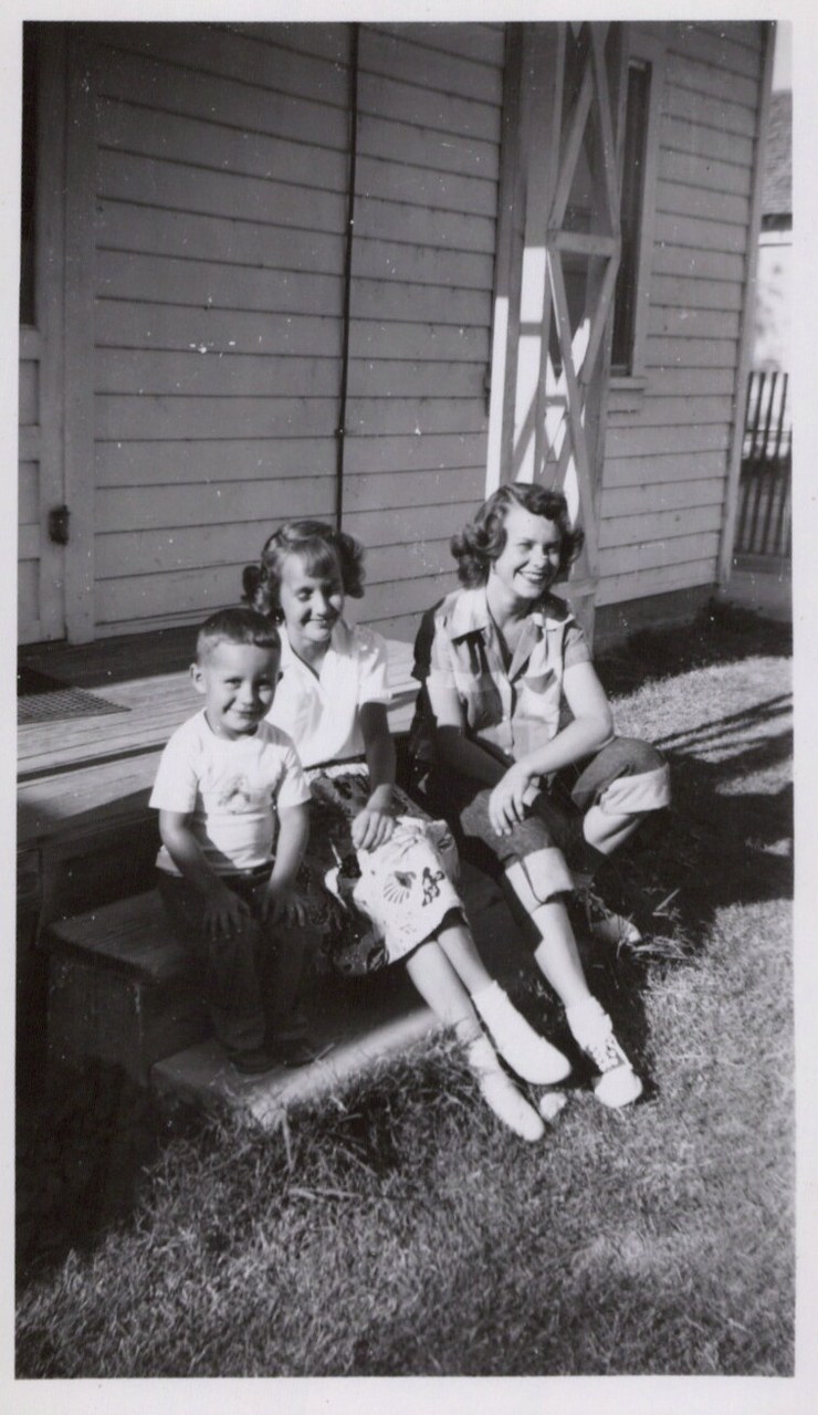 Three smiling children — a young boy and two girls — sitting on a step in front of a clapboard house, black-and-white photo, circa late 1940s–1950s