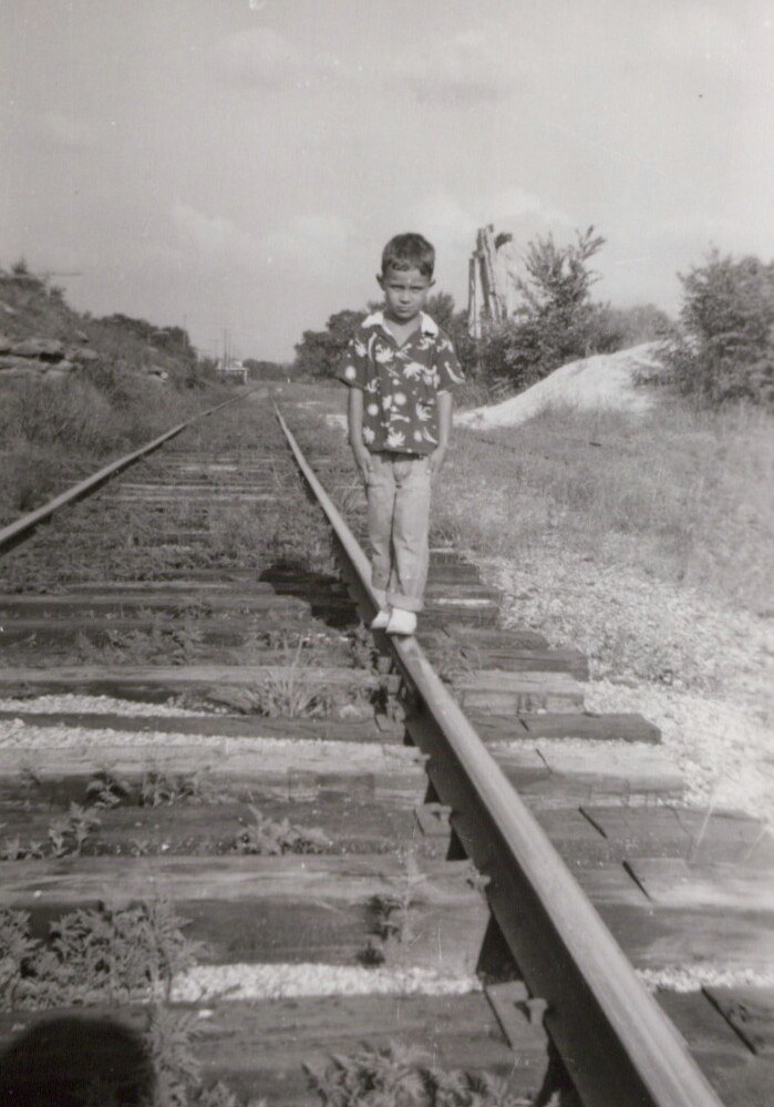 A young child balancing on a railroad rail and walking toward the camera on a lightly used rural track, wearing a floral shirt and light trousers, in a black-and-white photograph.
