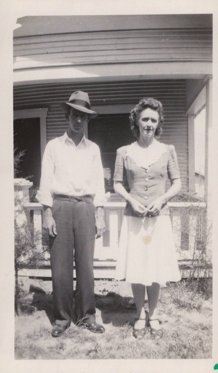 A man in a fedora hat and a woman in a button-front dress stand together in front of a wooden clapboard house in a black-and-white snapshot, likely from the 1940s.