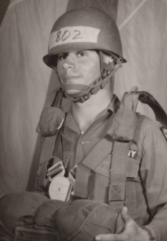 A young male soldier in a steel helmet numbered "802" and full parachute harness, seated in a black-and-white photo, likely at a military airborne staging area.