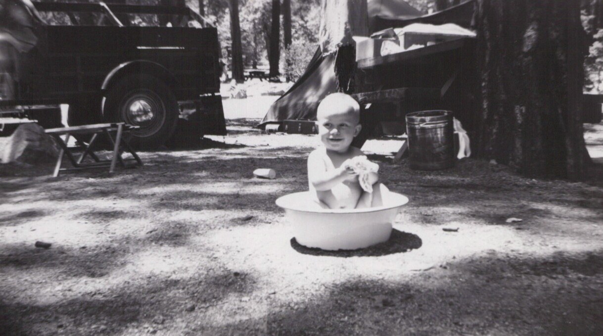 Smiling baby sitting in a round white wash basin outdoors at a forested campsite, with a vintage car and canvas tent visible in the background, black-and-white photo