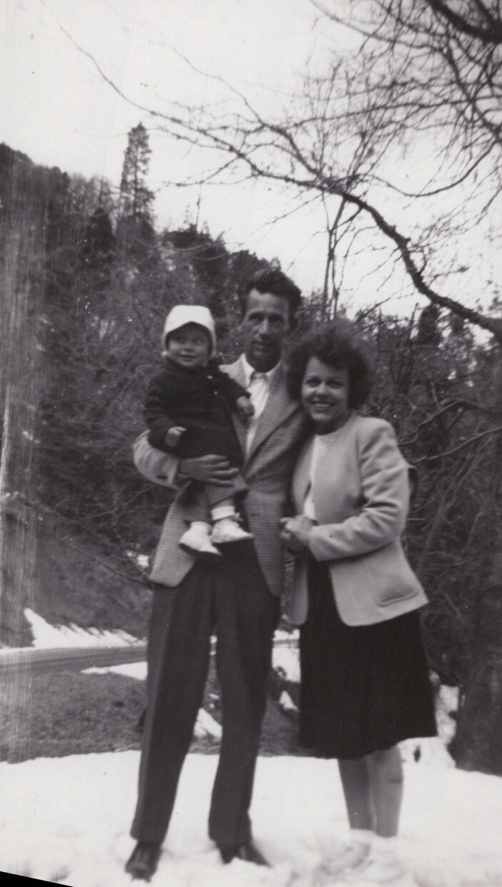 Black-and-white photo of a man holding a toddler in a white bonnet standing next to a smiling woman in a blazer, all posing outdoors in a snowy, wooded setting.