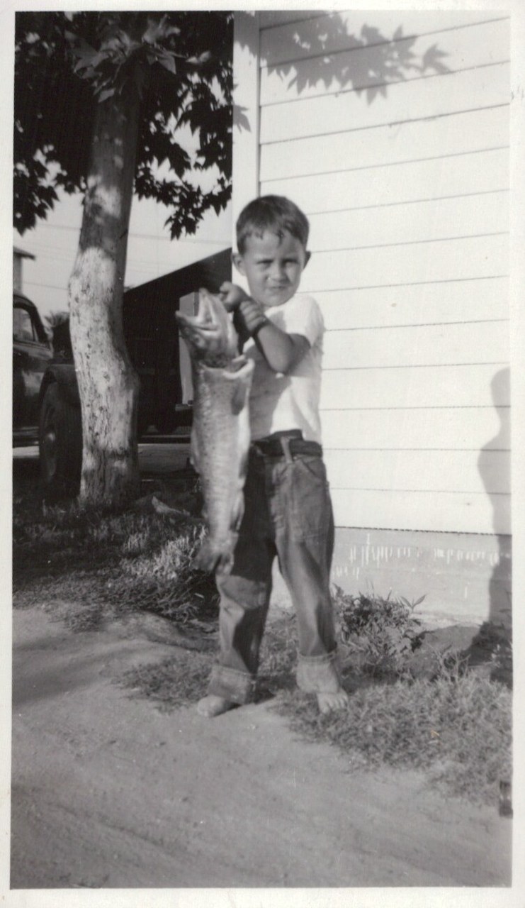 A barefoot young boy proudly holds up a very large fish in front of a white clapboard house, with a vintage car and tree visible in the background; black-and-white photo.