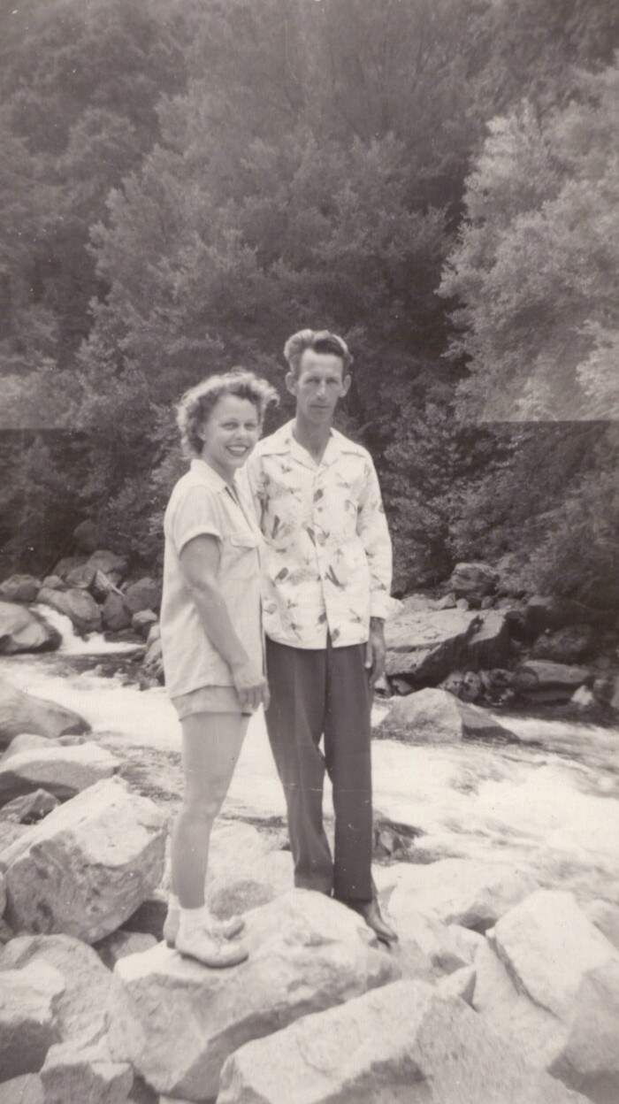 A smiling woman and a taller man stand on boulders beside a rushing mountain stream, with dense trees behind them, in a vintage black-and-white photograph.