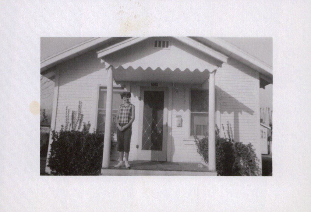 A woman in a plaid blouse and shorts stands on the front porch of a small white wood-frame house with a scalloped porch valance, in a black-and-white snapshot.
