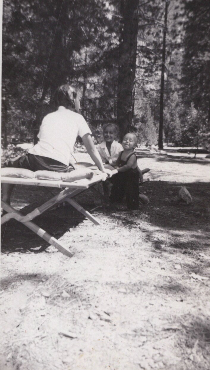 Black-and-white photo of an adult seated on a folding camp cot with two young smiling children in a shaded pine forest clearing