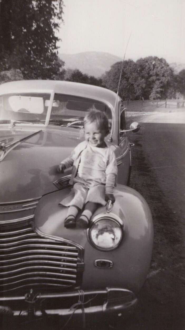 Smiling toddler sitting on the hood of a vintage 1930s–40s American car, with trees and hills in the background, black-and-white photo