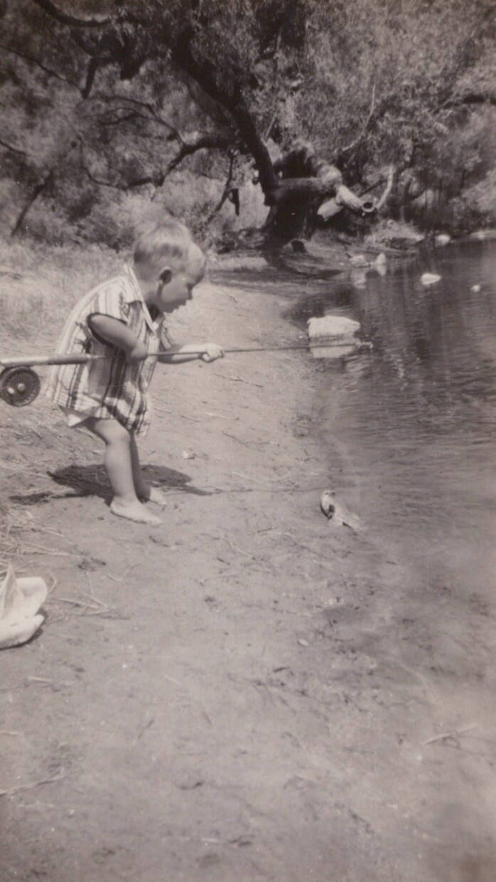 A barefoot toddler in a plaid romper holds a fishing rod on a sandy riverbank, with trees and calm water in the background, in a vintage black-and-white photo.