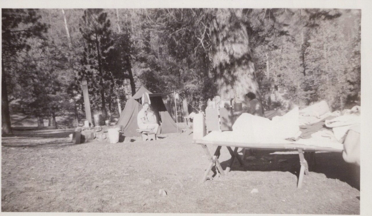 Black-and-white photo of a canvas tent campsite in a pine forest, with a person seated near the tent and camp gear on a folding table in the foreground