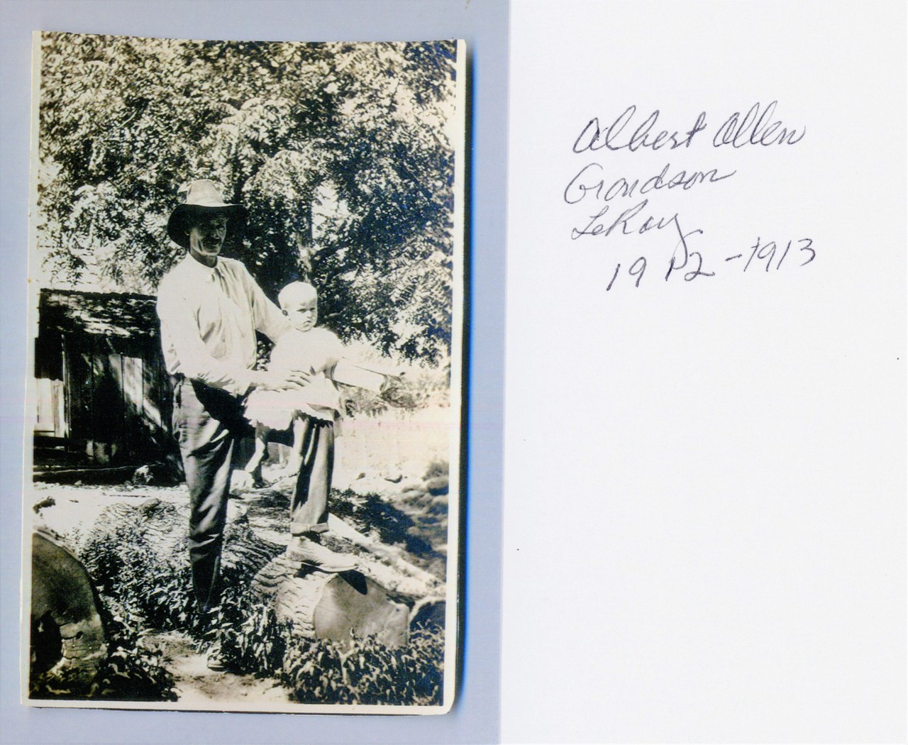 Black-and-white early 20th-century photo of an adult man in a wide-brimmed hat holding an infant outdoors near a rustic wooden shed, with a handwritten annotation card reading "Albert Allen / Grandson / LeRoy / 1912–1913" visible to the right.
