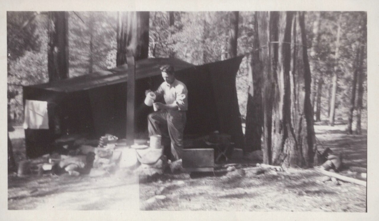 A man in a long-sleeve shirt pours from a camp vessel at a rustic woodland campsite sheltered by a dark canvas tarp strung between tall conifers, with scattered camp gear on the forest floor.