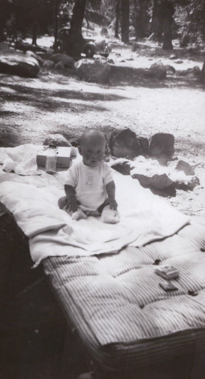 A baby in a white onesie sits on a striped mattress outdoors at a forested campsite in a vintage black-and-white photograph.
