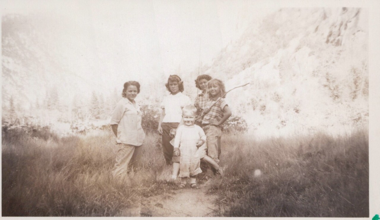 A sepia snapshot of an adult woman and four children posing on a dirt path in a canyon or mountain valley, with steep rocky cliffs and dry grass in the background.