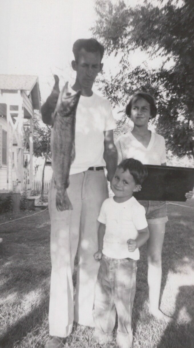 A young man holds up a large fish in a residential backyard while a smiling young child stands in front of him and a teenage girl stands nearby, black-and-white photo circa 1940s–1950s.