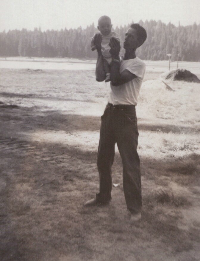A man in a white t-shirt and dark trousers holds a baby up at arm's length outdoors in a field, with a coniferous treeline in the background, in a vintage black-and-white photo.